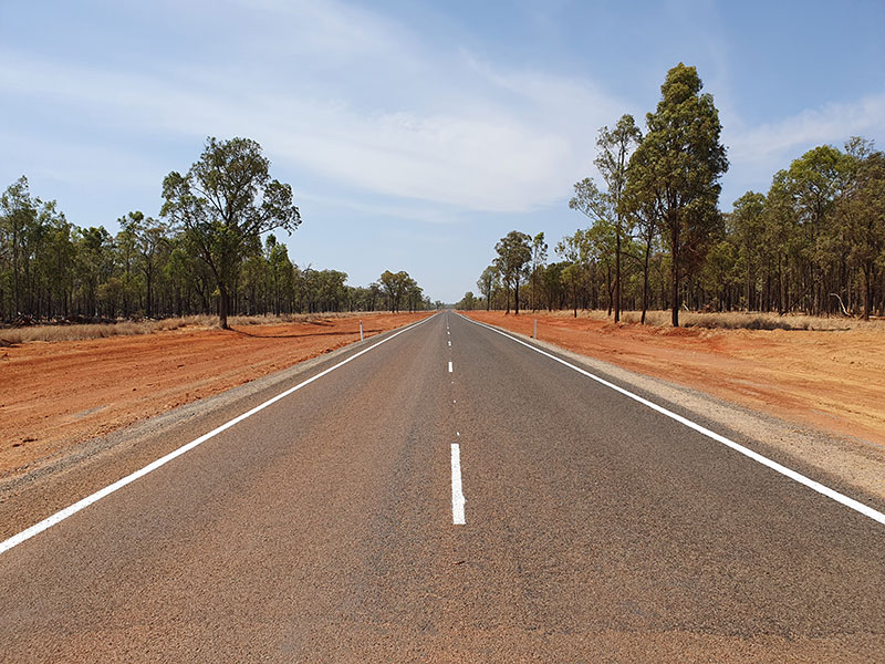 Camera looking face-on to to a road going into the horizon—red dirt either side of the road with trees.
