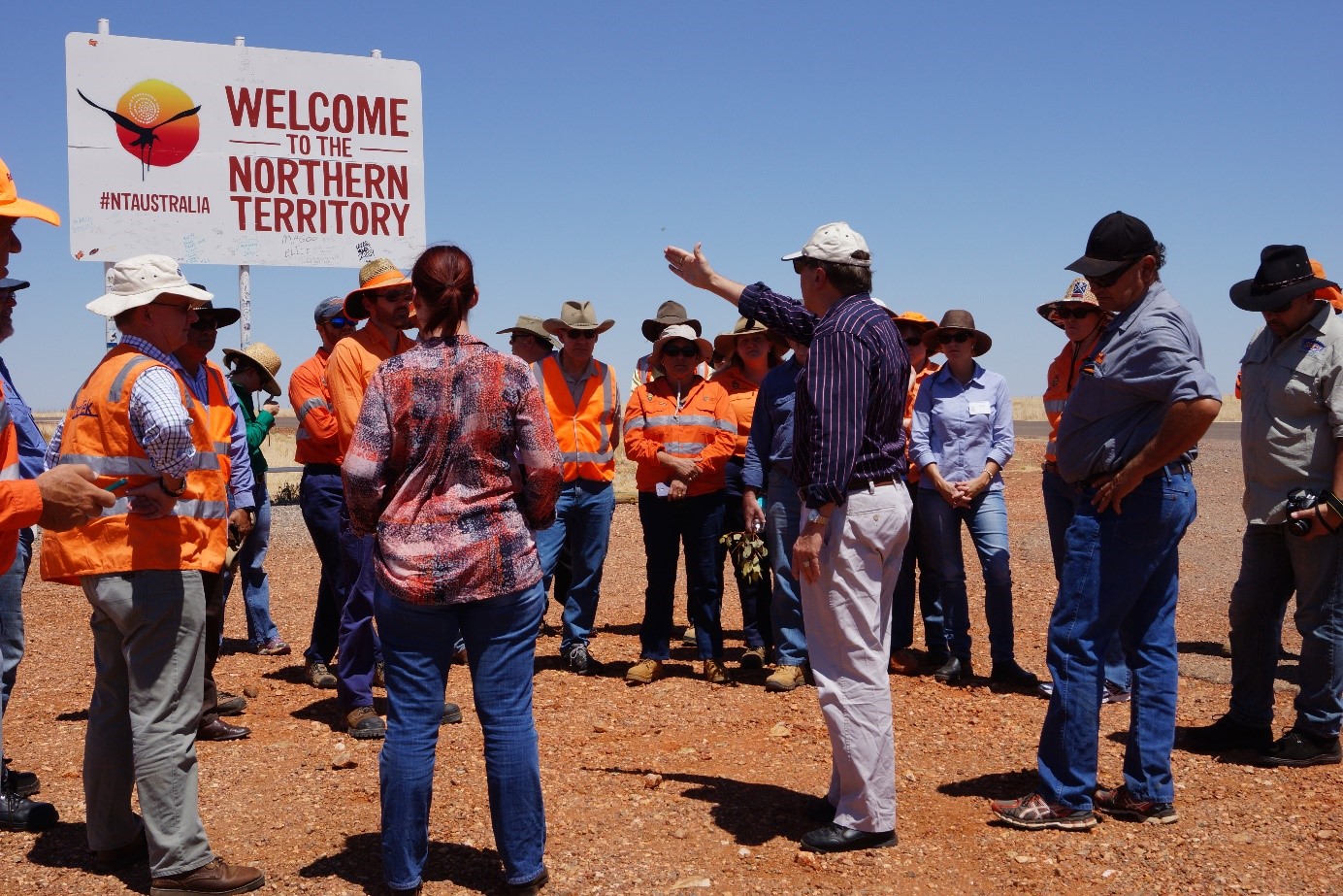 A person speaking to a group of people in high-vis clothing next to a 'Welcome to the Northern Territory' sign