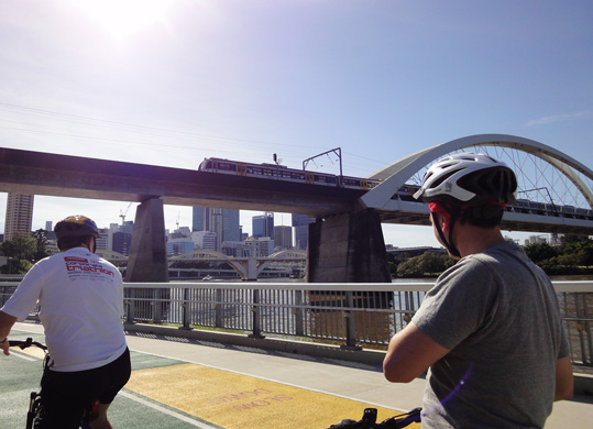 Image of two cyclists watching, a train crossing the Brisbane River on the Merivale Bridge, from the Bicentennial Bikeway.