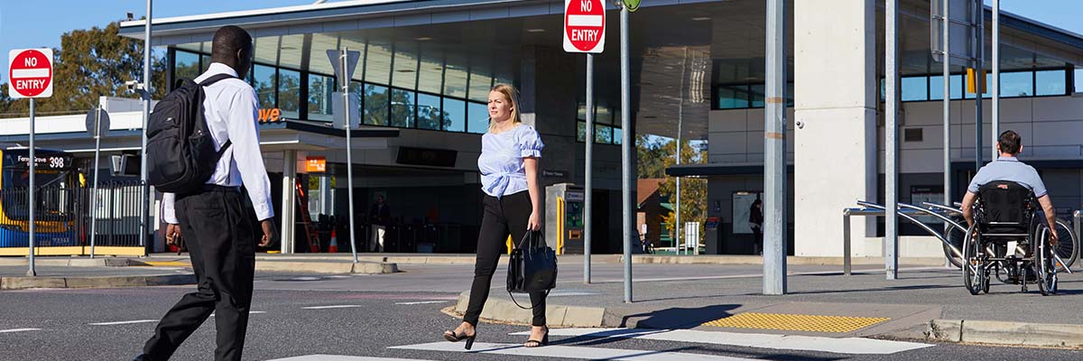 2 people dressed in corporate attire walking across a pedestrian crossing. A third person in a wheelchair is on the path. In the background a bus is stopped at a public transport facility.