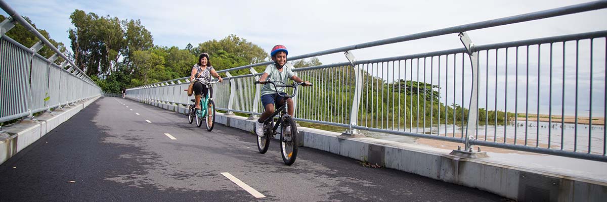 An adult and a child riding across a bridge. There is a waterway and trees in the background.