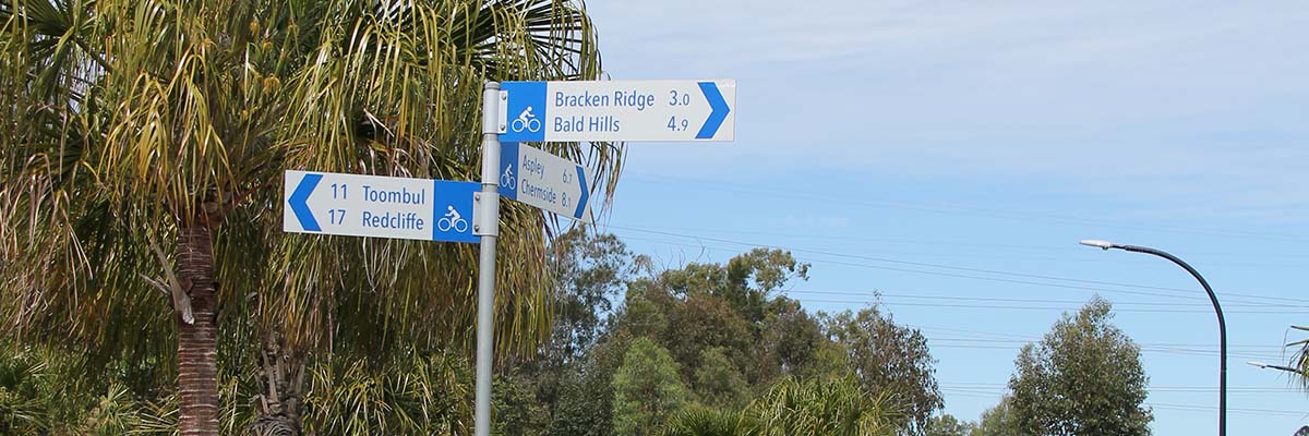 A street sign indicating direction and distances on a bike path. Toombul, 11. Redcliffe 17. Bracken Ridge, 3.0. Bald Hills 4.9.