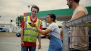 A woman showing her phone screen to a man in a hi-visibility vest, in the parking area of a service station.