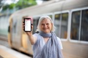 A woman standing in front of a train, holding her phone showing her Digital Licence, to the camera.