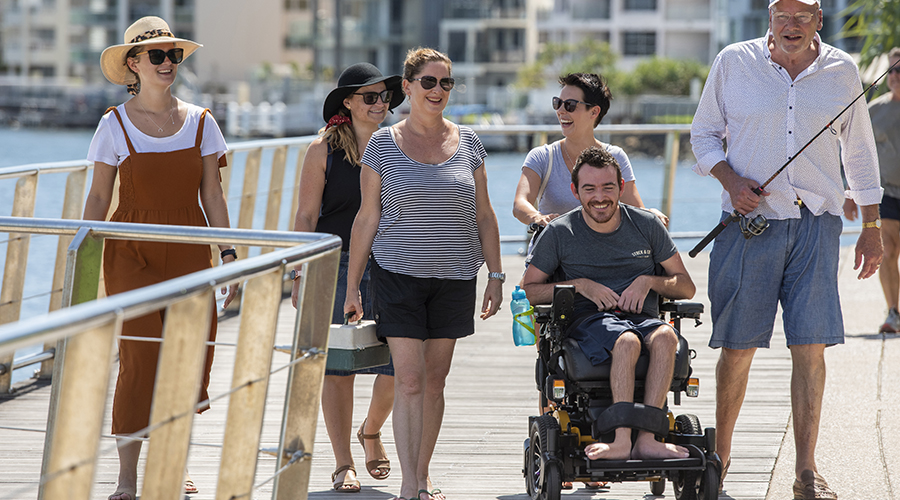 Group of friends walking along accessible walkway at ocean. Image includes women, senior carrying a fishing rod and man in motorised wheelchair.