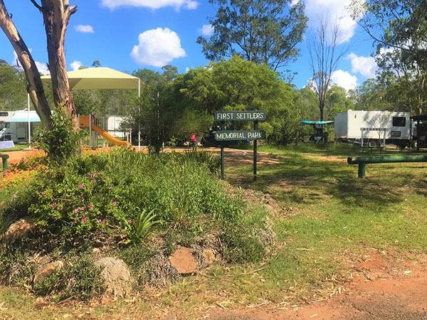 A tree with plant life in front of a playground and a sign that says First Settlers Memorial Park