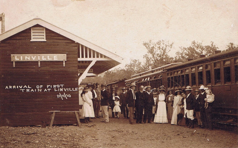 A sepia photo of people gathering at a train, with a sign that says 'Arrival of first train at Linville