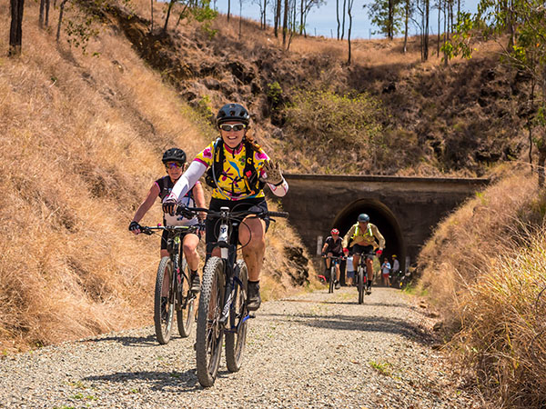 Bicycle riders emerging from a tunnel, with 2 in the foreground smiling and waving to the camera
