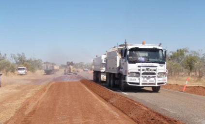 Photograph shows road construction in a dry rural area, with a white dump truck traveling along a newly surfaced lane beside reddish gravel. Dusty unpaved roadway, orange safety markers, earthmoving equipment, and sparse trees highlight active paving work under clear skies.
