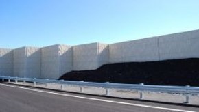 Photograph showing a divided highway beside a tall segmented concrete sound barrier or retaining wall. Bright blue sky, dark wall base, metal guardrails, and multiple roadway lanes create a clear transportation-infrastructure scene.
