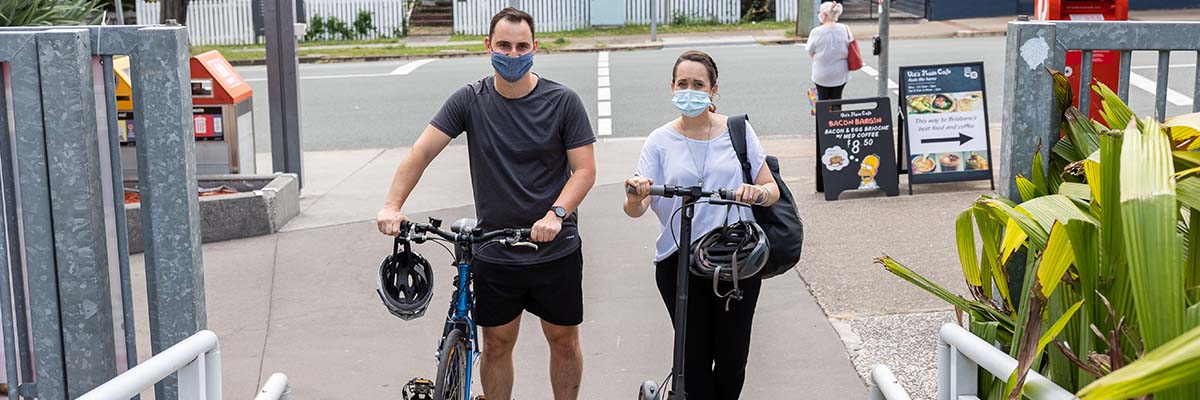 2 people standing together. One stands with a bike, the other with a scooter.