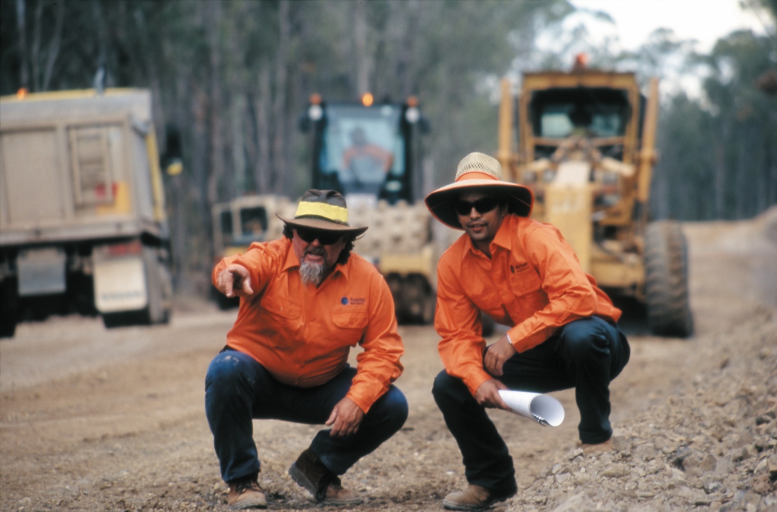 Two people in high vis squatting in front of road plant