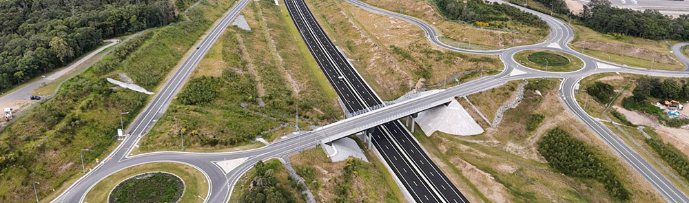 View looking south at the completed Flood Road interchange on the Bruce Highway Cooroy to Curra Section D, featuring roundabouts, overpass, on/off ramps, new pavement, and Noosa Road bridge in the distance.