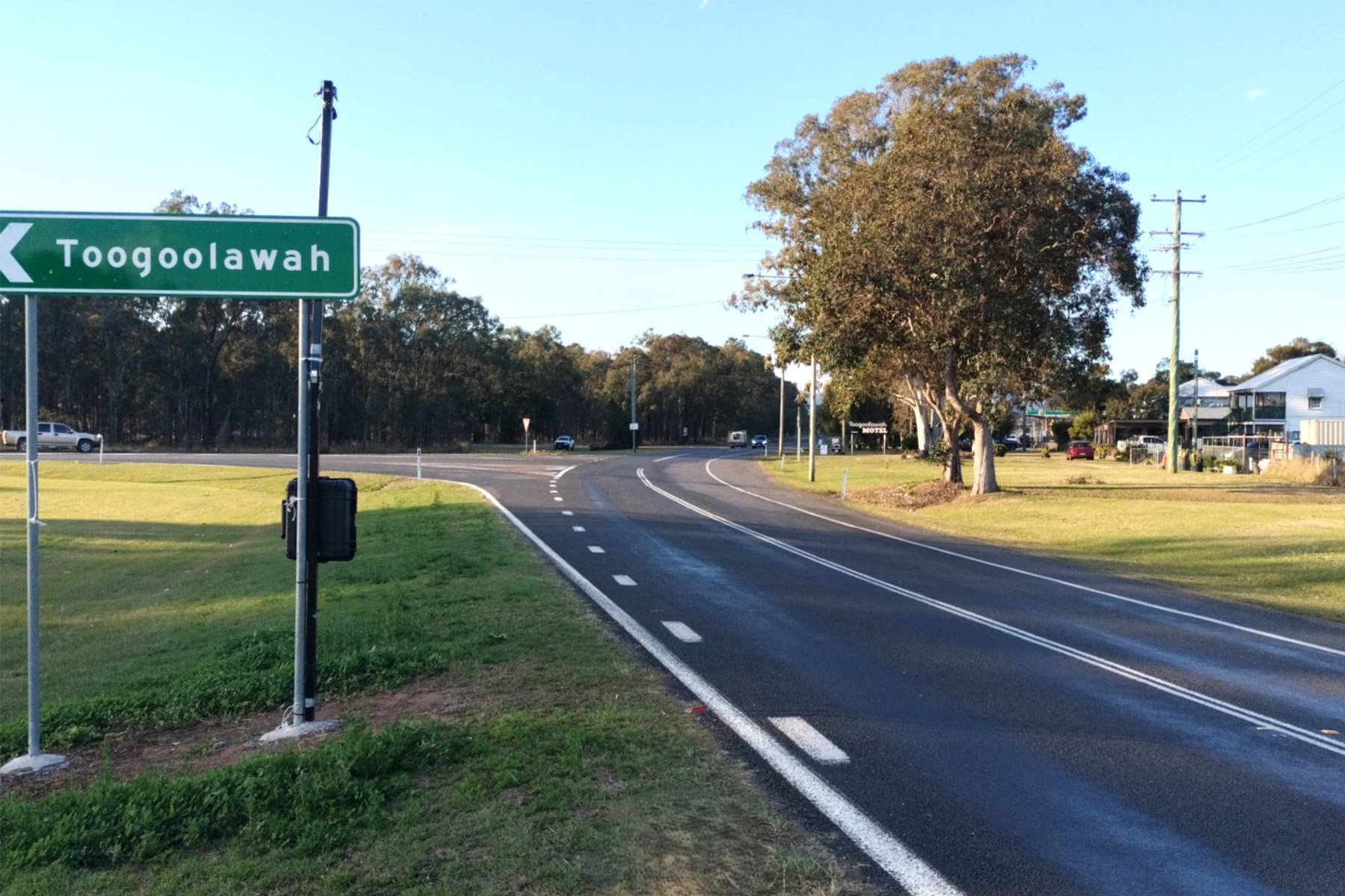 Road with a green sign with an arrow and Toogoolawah in white writing and trees and building on the right hand side of the image.