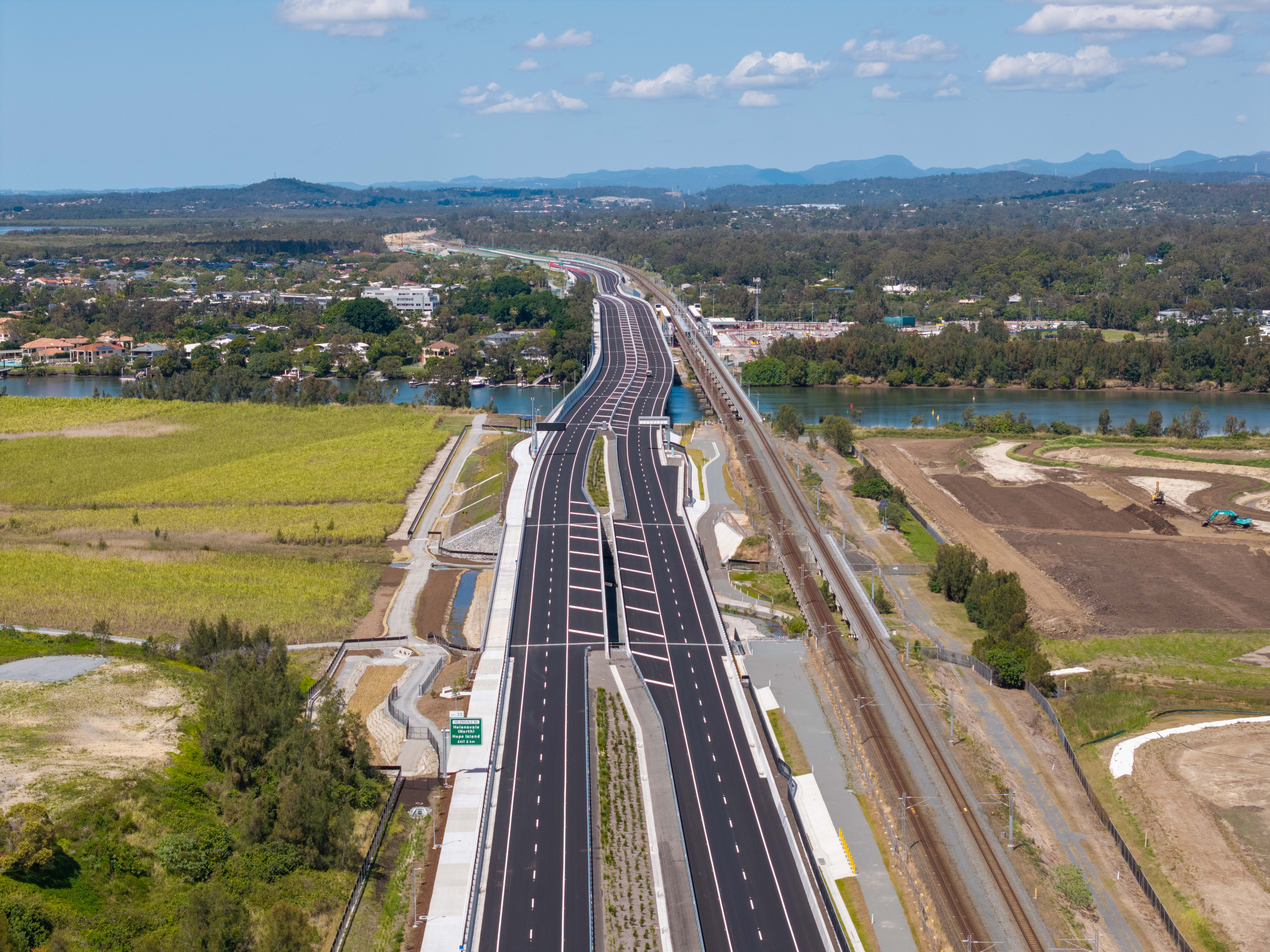 Ariel view of the Coomera Connector highway looking south over Nerang River with paddocks on either side of the highway, residential area across the river with mountain in the background. 