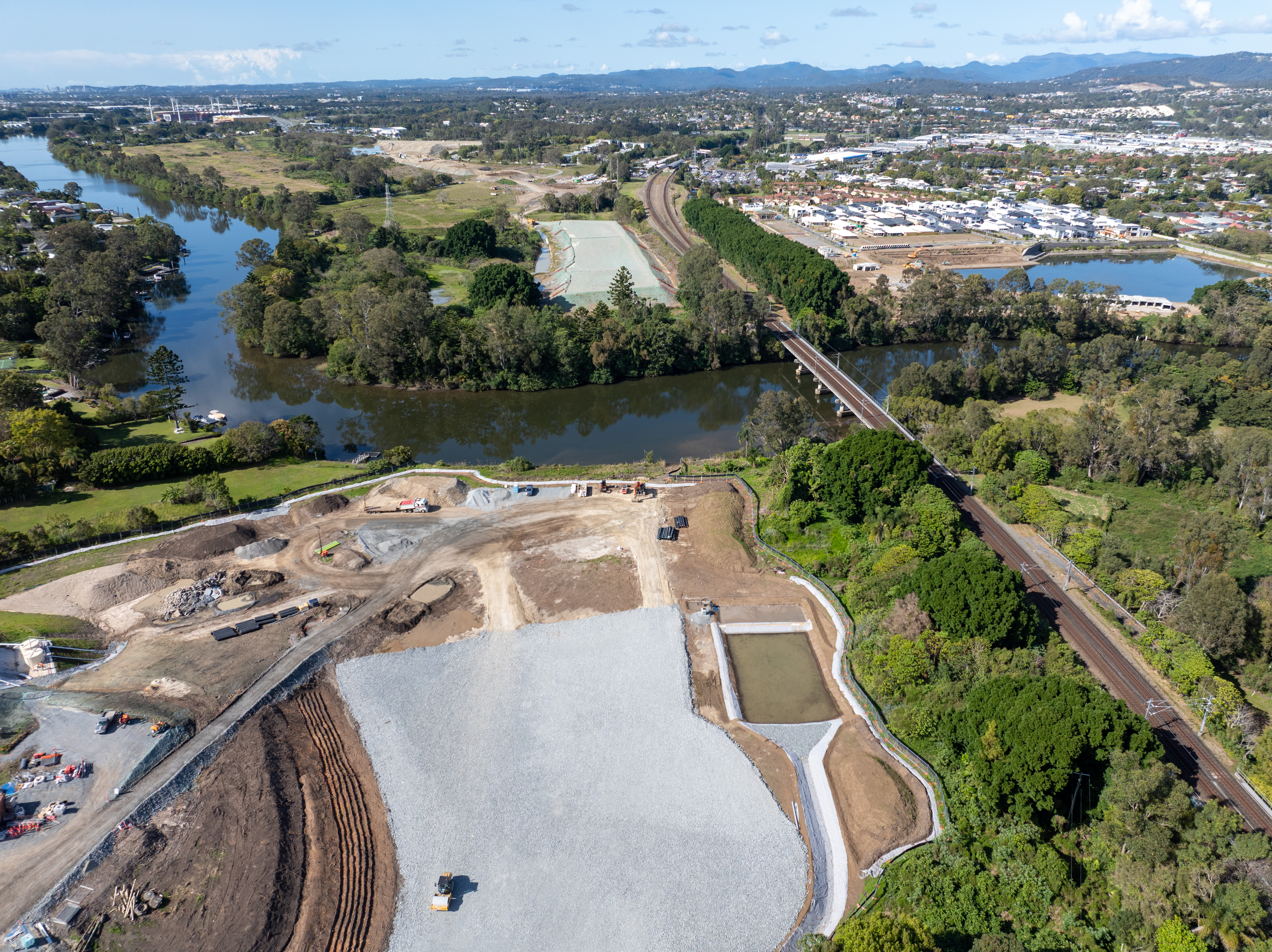 Construction site next to Nerang River with train tracks on the right-hand side of the site and building across the river with trees along the river and next to the site.