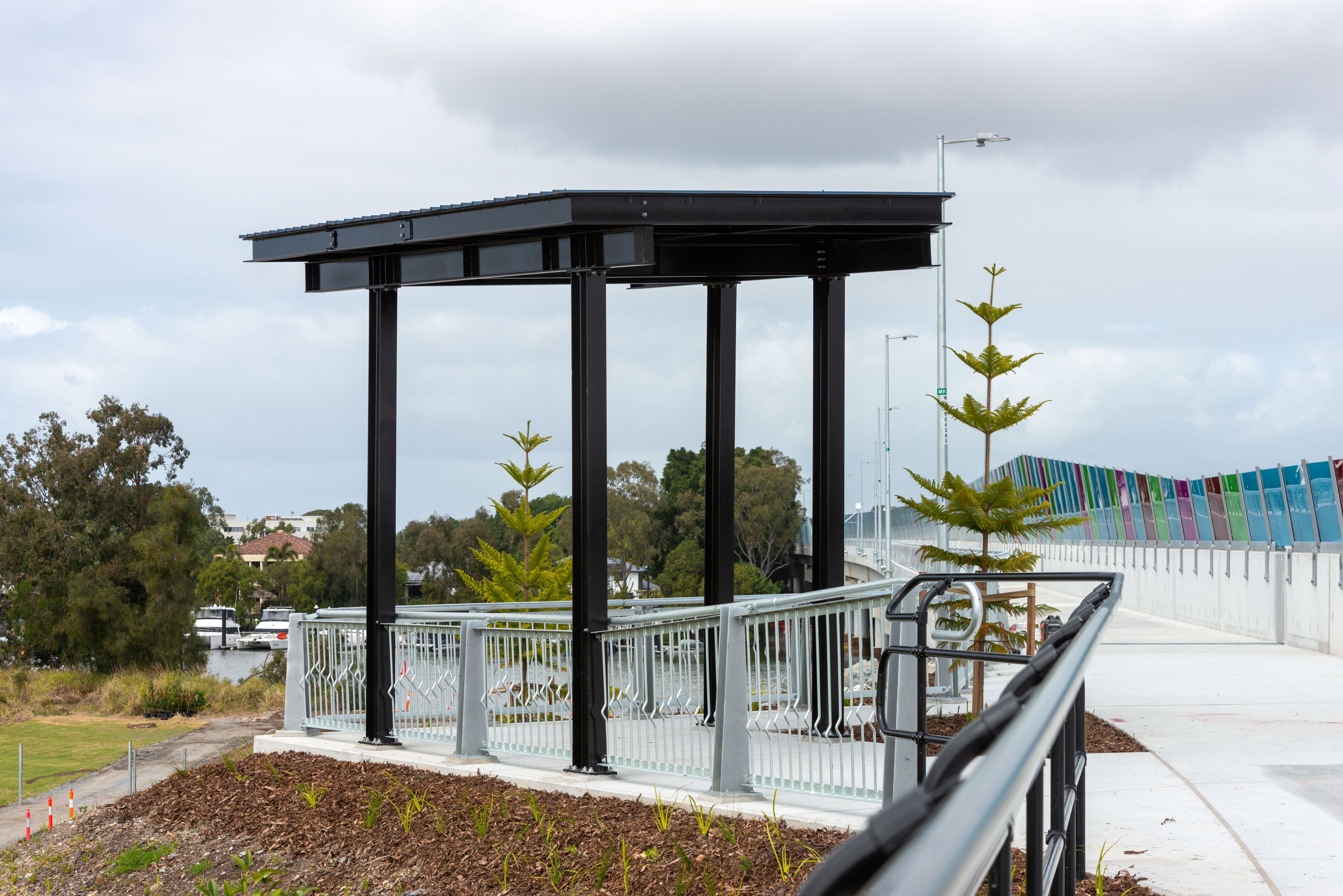 A black covered rest area at the end of a walking and bike path at Coomera. The black cover as four poles and a roof and it is off to the left-hand side with a railing around the front and side of the rest area.