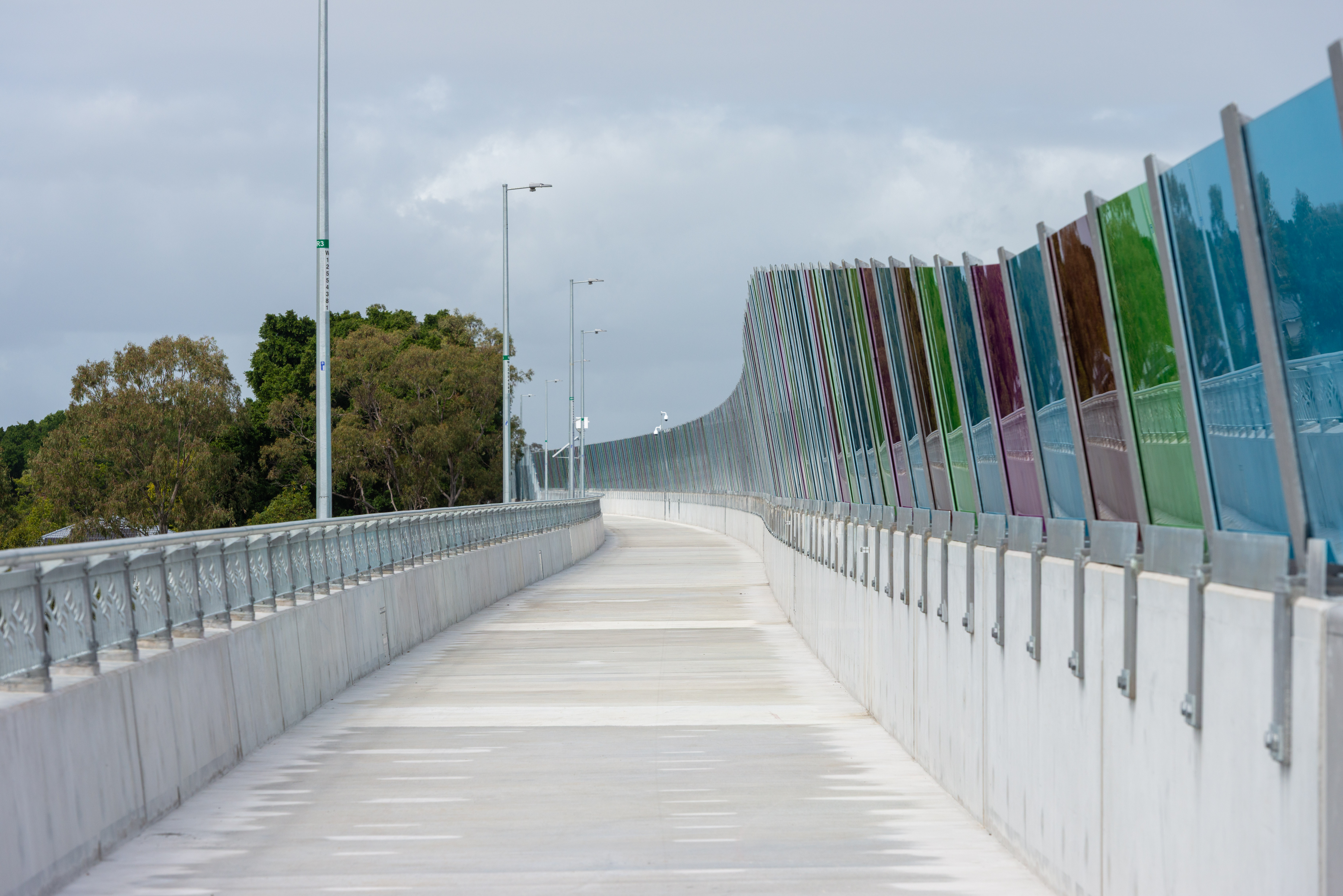A pathed walk way with blue, green and red coloured barrier on the right-hand side with trees and light poles on the left.