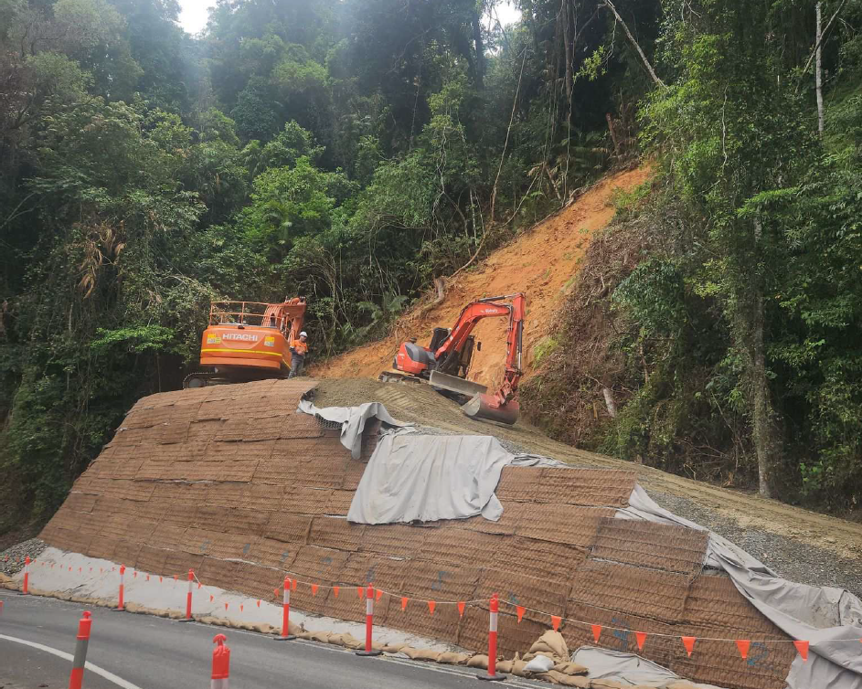 Construction machines on side of a hill and road building stability of the upslope on Mossman - Mount Molloy road with trees on either side of the site. 