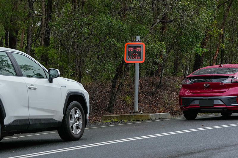 Roadside sign displaying a red 'Too close' message in response to a white vehicle travelling too close behind a red vehicle.