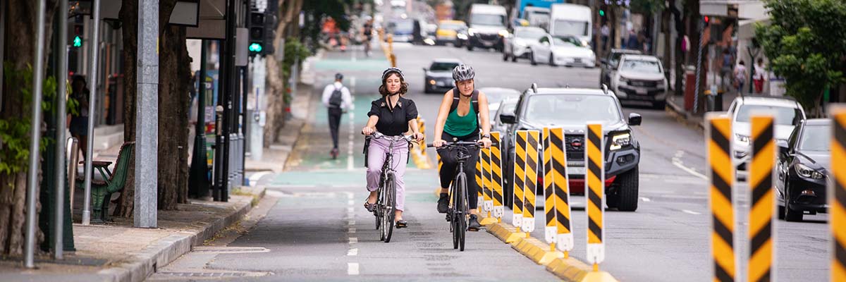 2 people riding bikes on a separated bike path adjacent to a busy urban road.