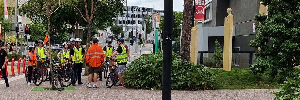 A group of people wearing high viz vests, standing with bikes on a pathway by a city road