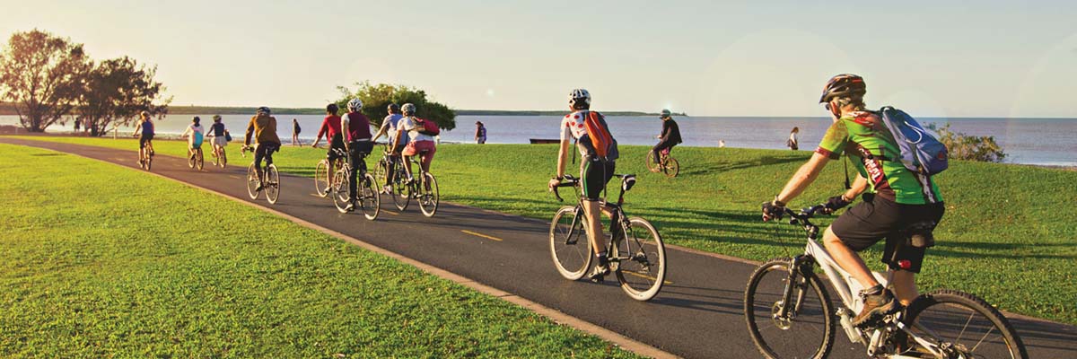 A group of people riding bikes on a shared path with a park and ocean in the background.