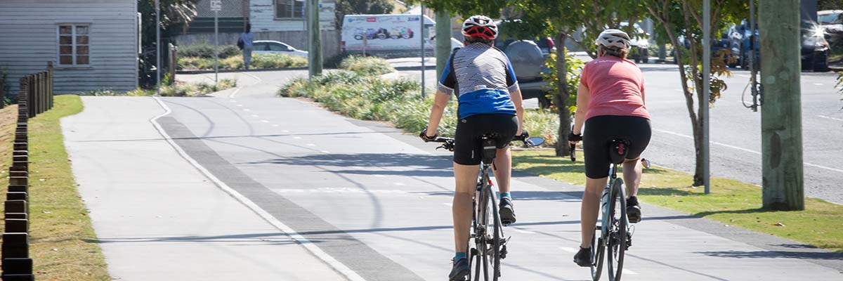2 people riding bikes along a shared path near a road.