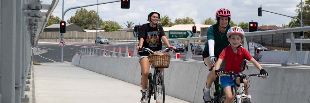 A child, and adult and a teenager ride bikes on a path next to an outer-city road. All 3 wear helmets and the child and adult wear gloves.