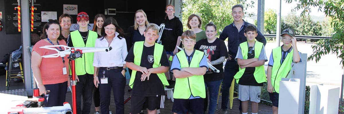 A group of adults and young teens stand in a covered outdoor space smiling at the camera. Some wear safety glasses and some of the young teens wear high viz vests. In the foreground part of a bike frame is held up on a metal stand.