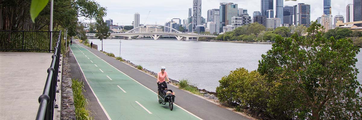A person riding an adapted bike with a storage compartment at the front along a path beside a river, with a city skyline on the other side of the river.