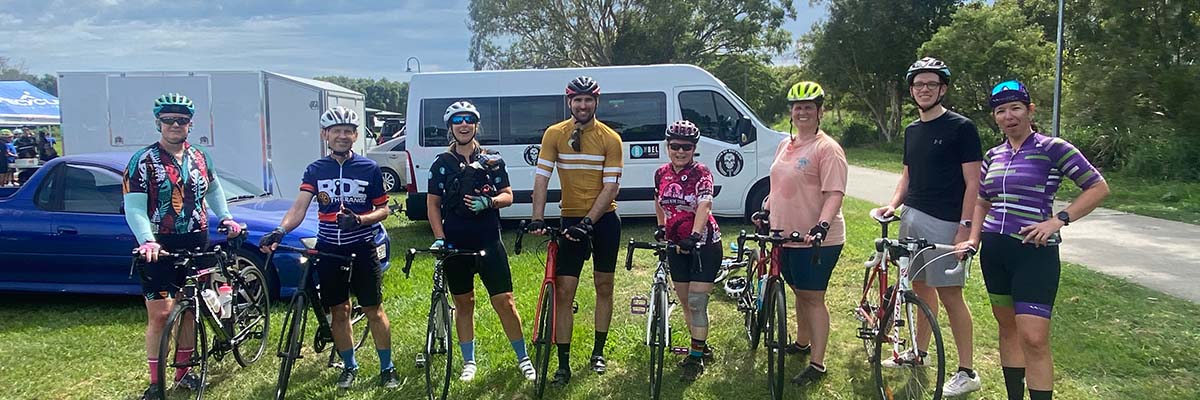 8 people in bike riding attire stand with 7 bikes at a grassy outdoor space.