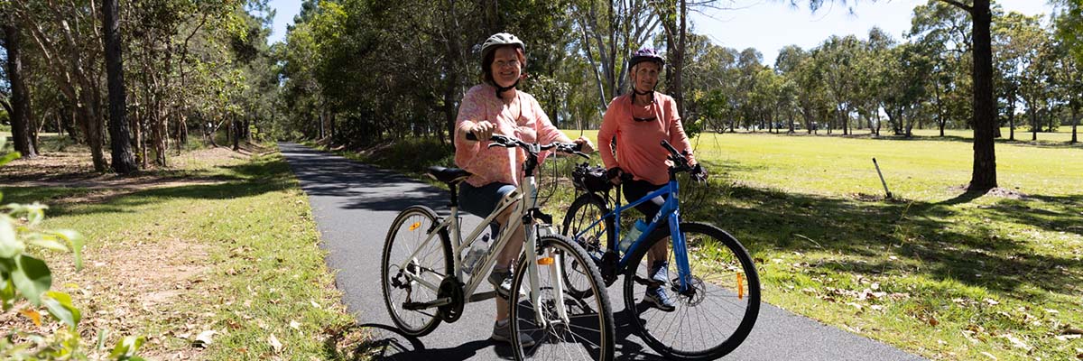 2 middle-aged women stand with bikes on a tree-lined path.
