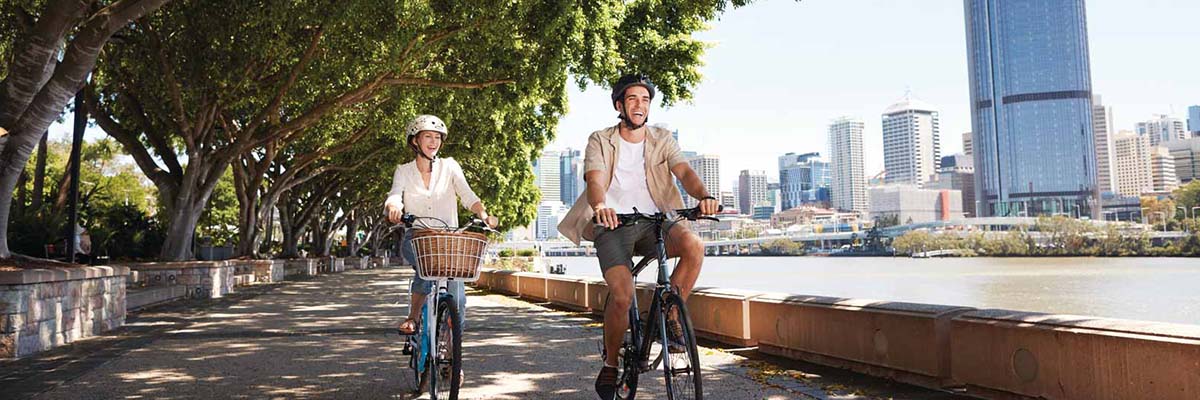 2 people ride bikes on a shady tree-lined path beside a river, with a city skyline on the other side of the river.