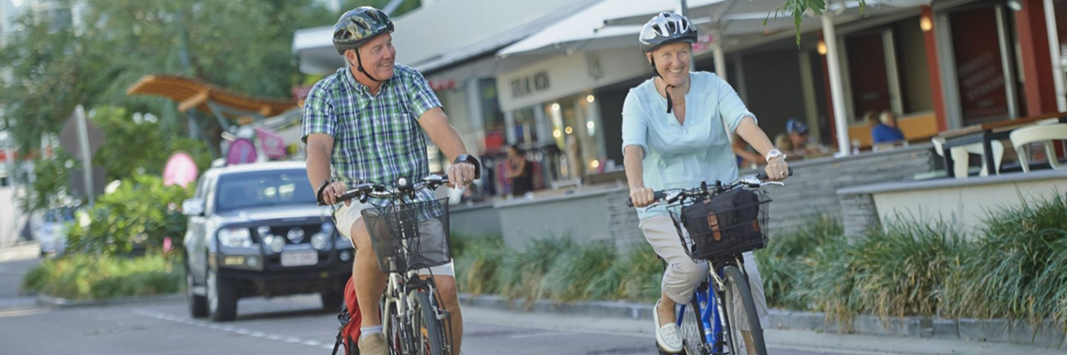 2 people riding bikes on a roadway in front of a row of store fronts and restaurants.