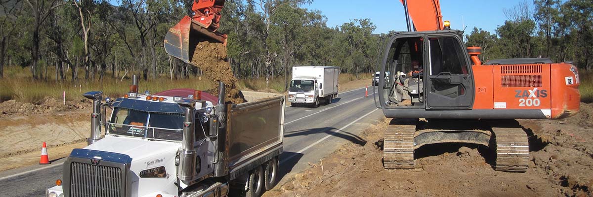 A digger on a large pile of dirt next to a road, pouring dirt into the back of a truck on the road.