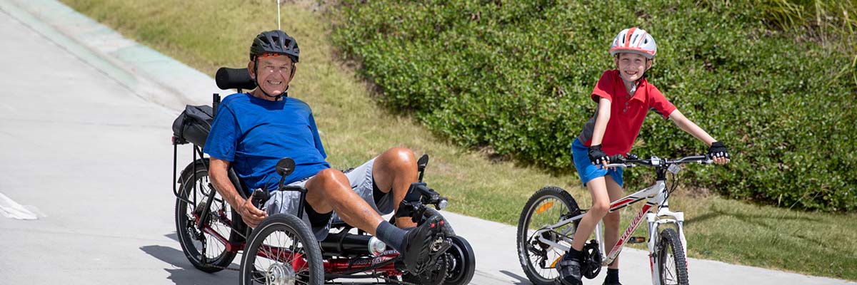 A child riding a bike next to an older person on a recumbent trike, on a downhill concrete path in a grassy area.