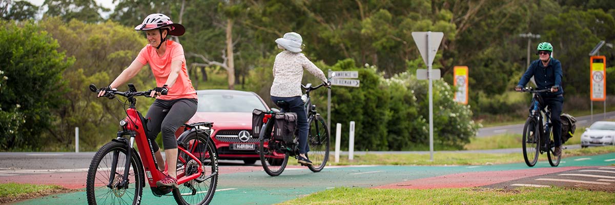 3 bike riders, 2 riding in one direction and one in the other, on a bike path running parallel to one road and intersecting with one other that turns off at a T-intersection. There is a speed bump on the road where it crosses the bike path, and 2 of the bike riders are crossing on it. A car is turning from the parallel road into the intersection.