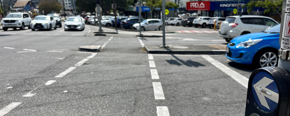 A intersection on a street in Queensland with traffic lights, cars and a pedestrian crossing. Nearby there are local businesses and high-rise buildings.