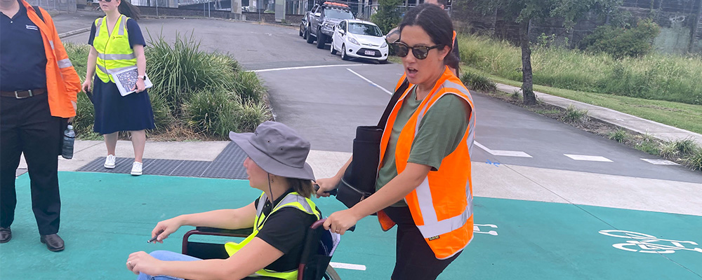 Woman in high visibility vest pushing a person in a wheelchair across a green path across a street.