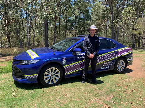 A travel inspector wearing a navy blue uniforms standing in front of a navy transport inspector vehicle with magenta, white and yellow decals