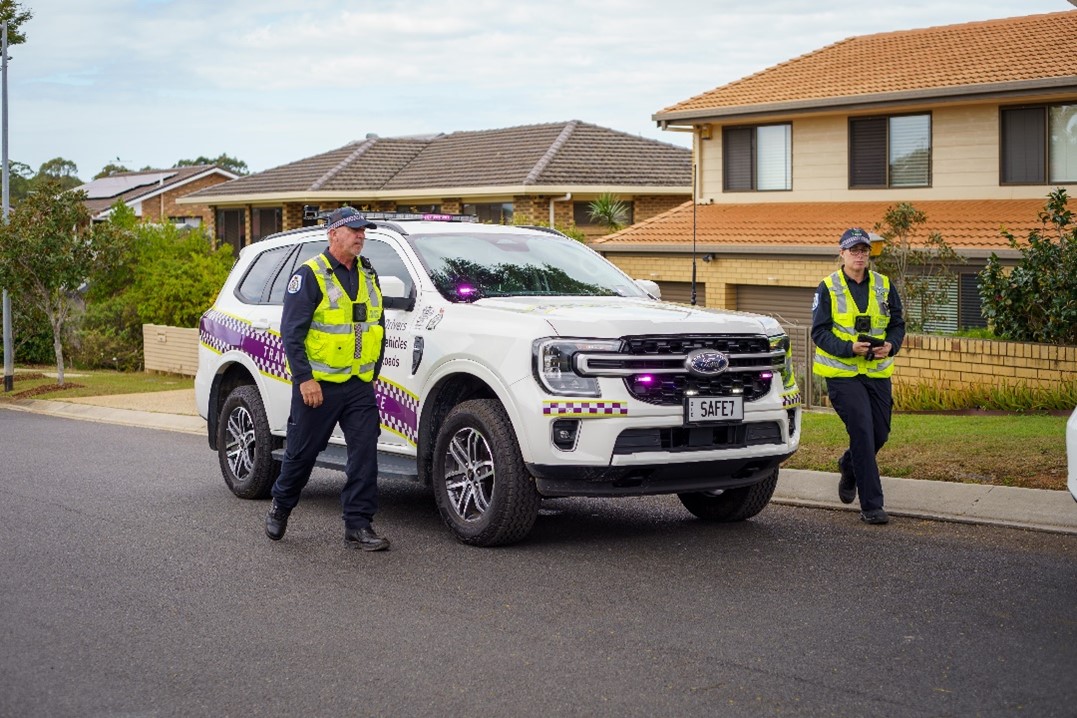 Transport inspectors with navy blue uniform and high visibility vests standing near a white vehicle with flashing lights. 